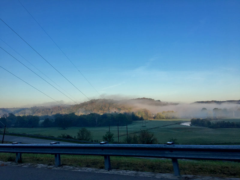 View from a roadside guardrail with green fields, tree and fog covered hills in the distance
