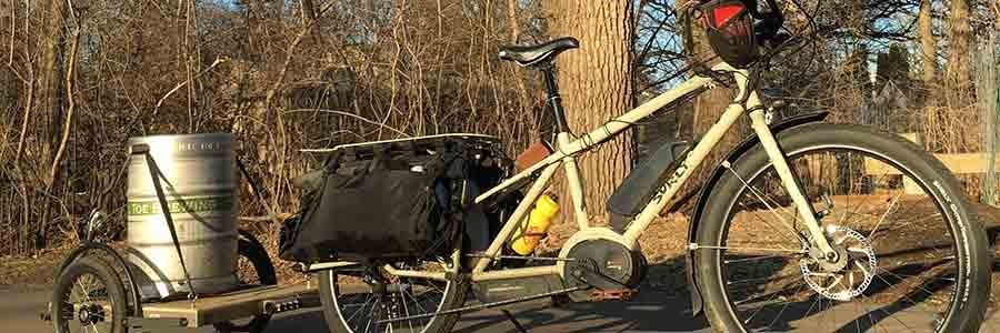 Right profile view of a tan Surly Big Easy bike with gear pulling a trailer with keg on a paved trail with trees behind