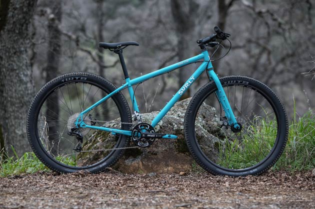 Right side profile of a Surly Bridge Club bike, turquoise, in front of a big rock and woods behind