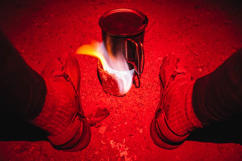 Downward view of a steel cup with a flame underneath, between 2 sandal clad feet, and red flashing beam shining over
