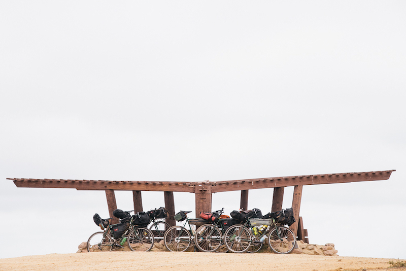 Side profiles of gear loaded bikes lined up in front of a wooden pergola on a sandy plot