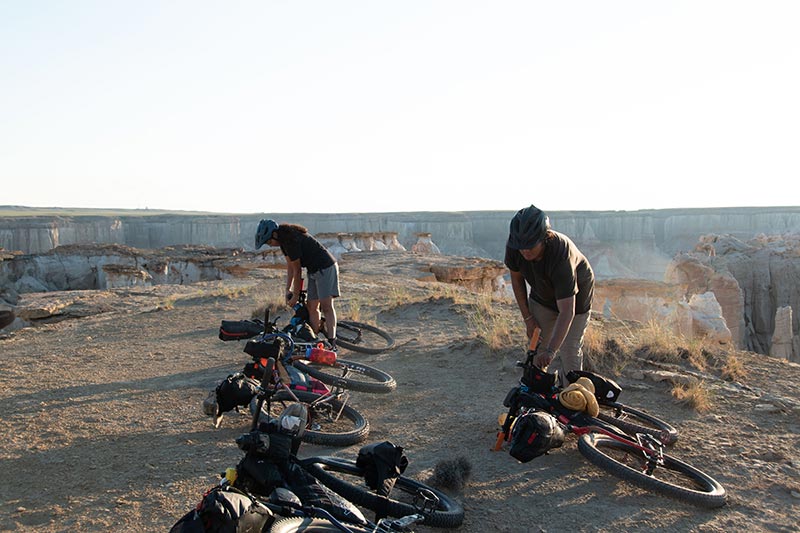Four loaded mountain bikes laying on ground, two people getting bikes ready to ride out after packing up camp