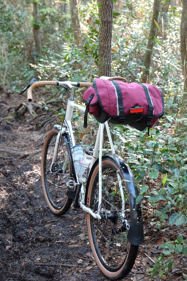 Surly Midnight Special bike, white, with red seat pack facing away down a dirt trail in the woods