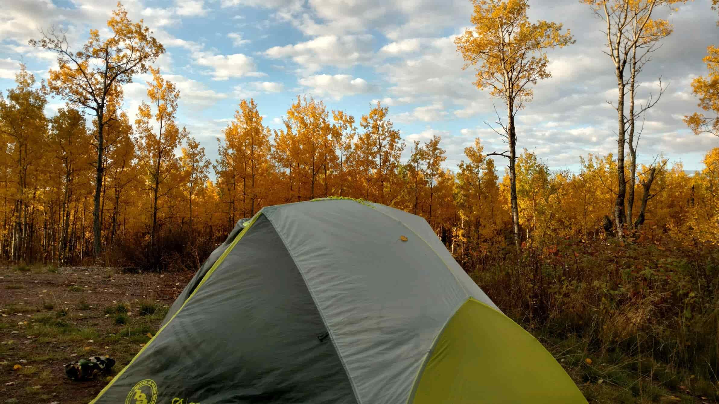 A gray and lime green dome tent on a grass field - trees with yellow leaves, blue sky and white clouds in the background