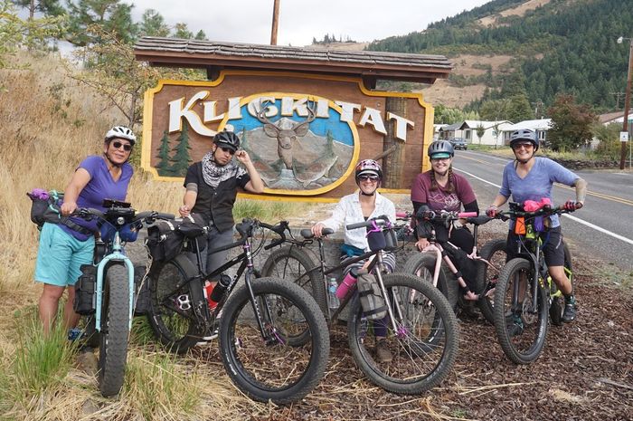 Group of 5 cyclists standing with their bikes with gear in front of a Kligkitat sign on the shoulder a mountain road
