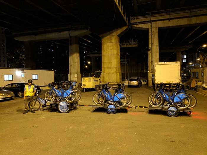 Person standing under a bridge at night in front of a three wheeled bike with two trailers loaded with bikes connected